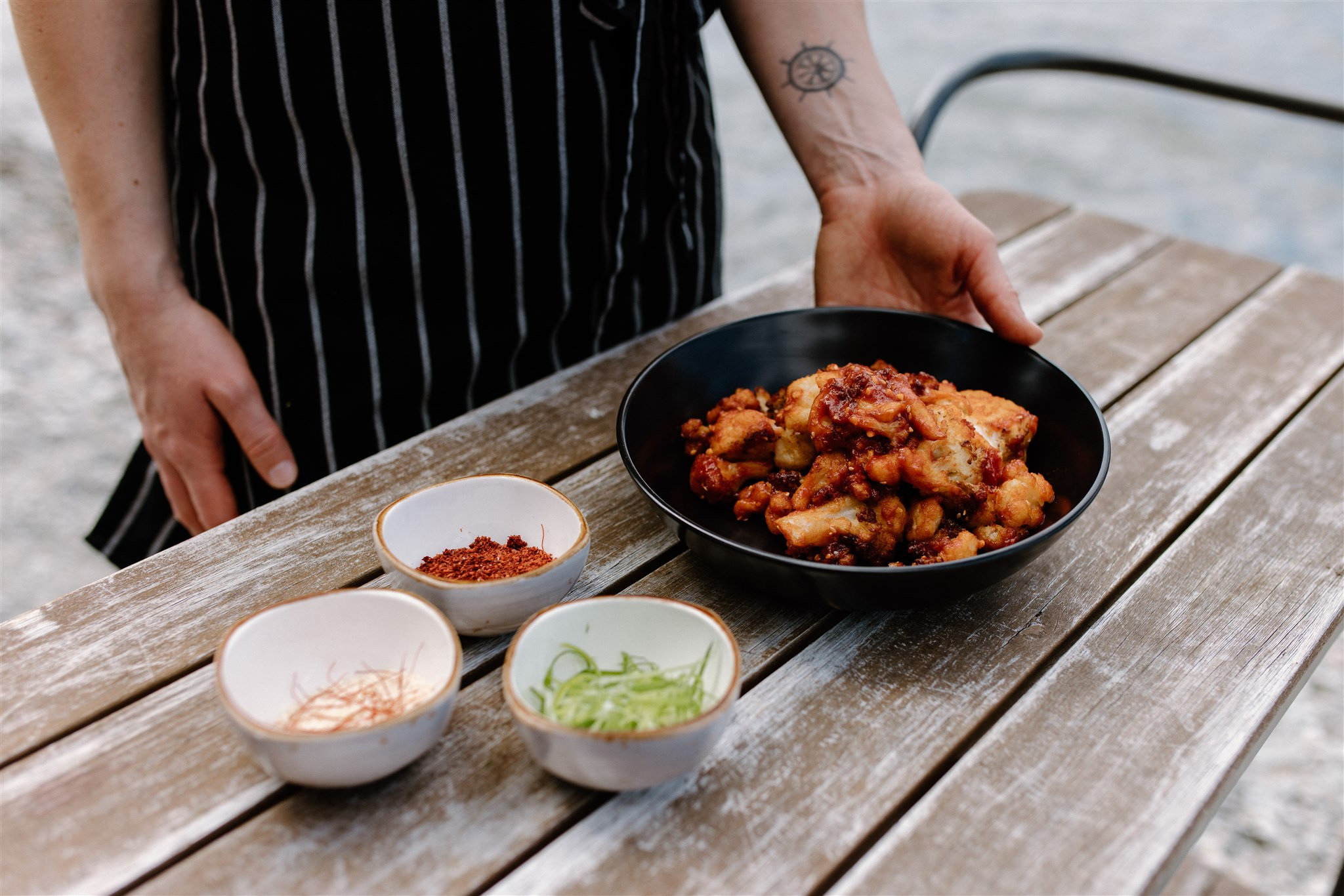 A close up of someone setting down a bowl of our Korean Cauliflower next two three little dishes on spices, on a wooden table next to the lake.