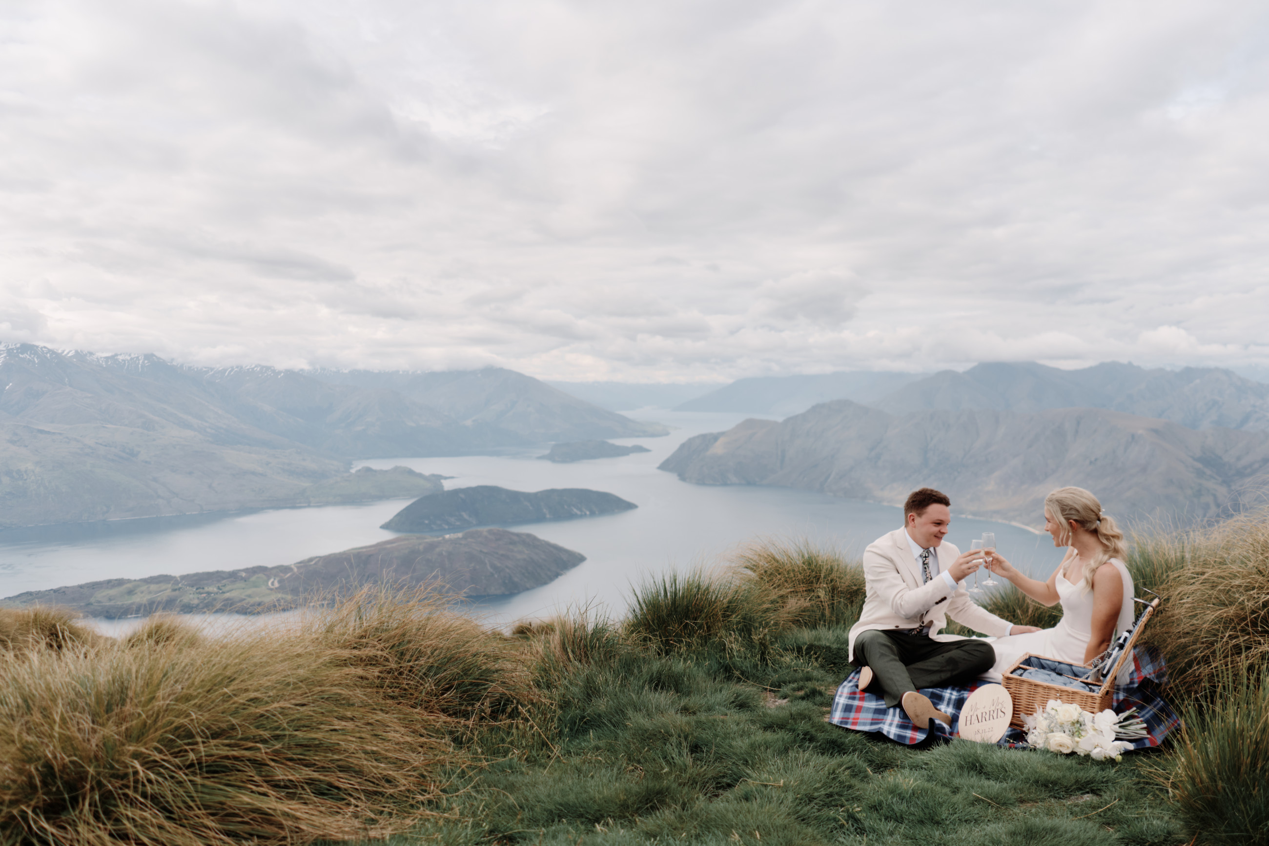 Couple sitting with a picnic hamper cheersing at the top of Coromandel peak. Lake Wānaka in the background.