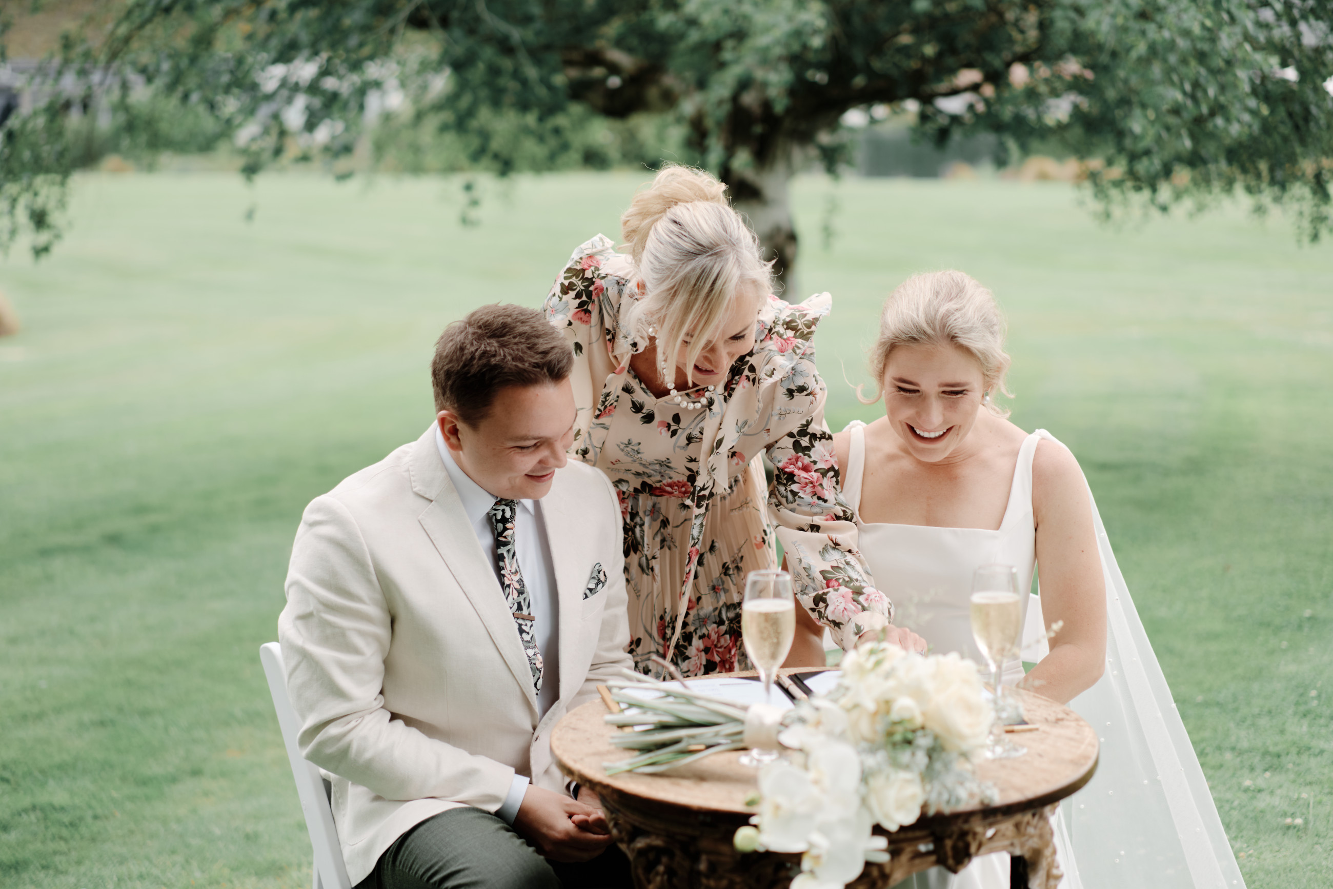 Couple and their celebrant laughing at the signing table. Two glasses of Champaign and bouquet of flowers sitting on table. Grassy lawns and a tree in the background