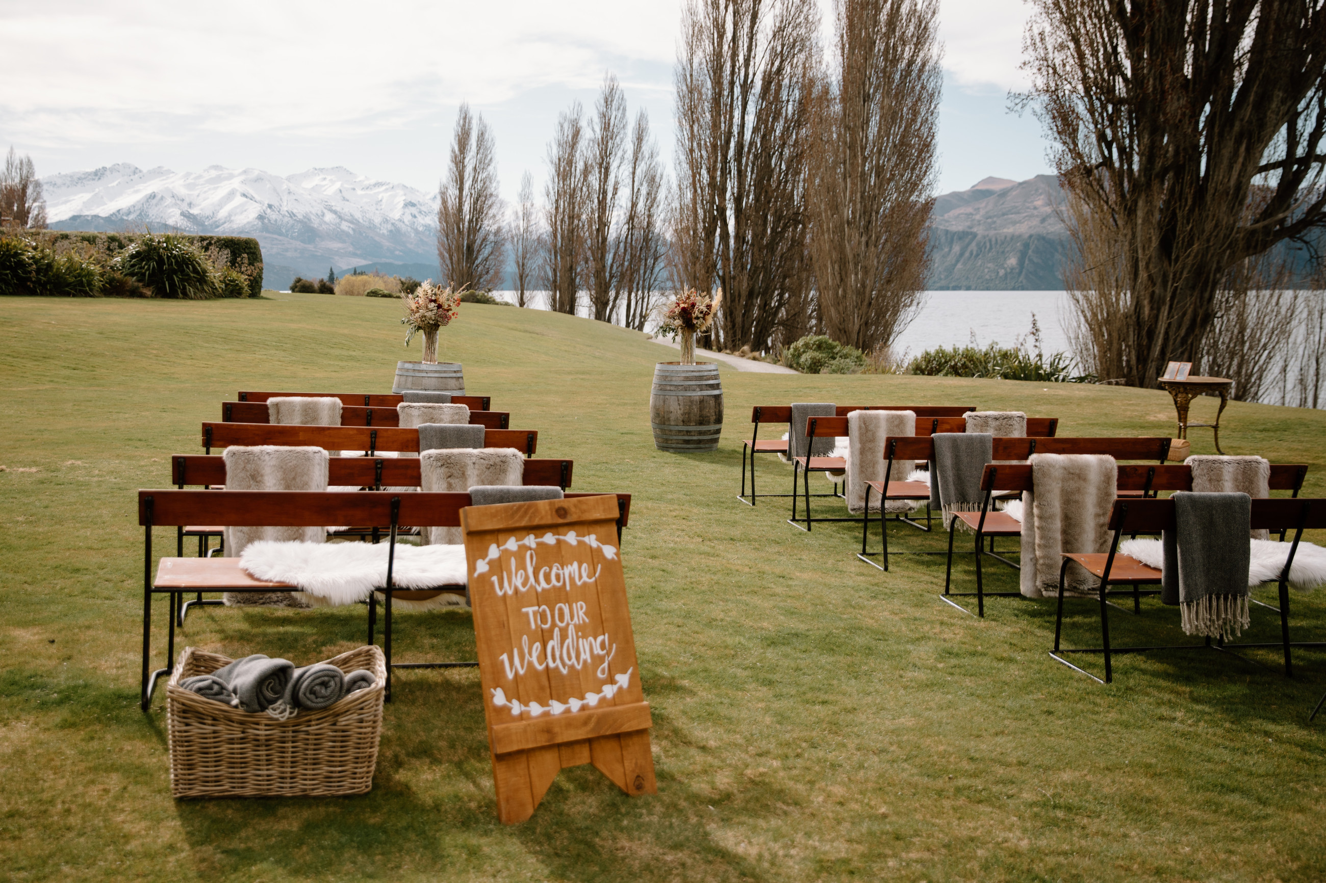 A wedding ceremony set up with brown bench chairs draped with rugs and sheepskins. In the background are rolling green hills, snow caped mountain and Lake Wānaka