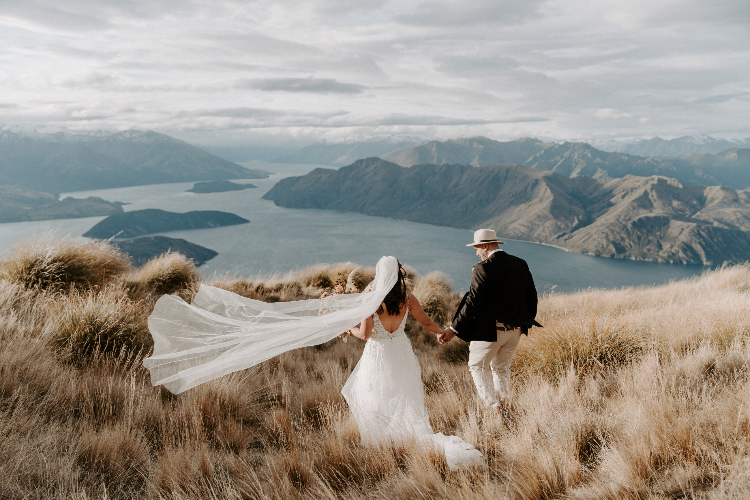 Wedding couple walking through tussoks at the top of coromandel peak. Stunning view of Lake Wānaka in the background.