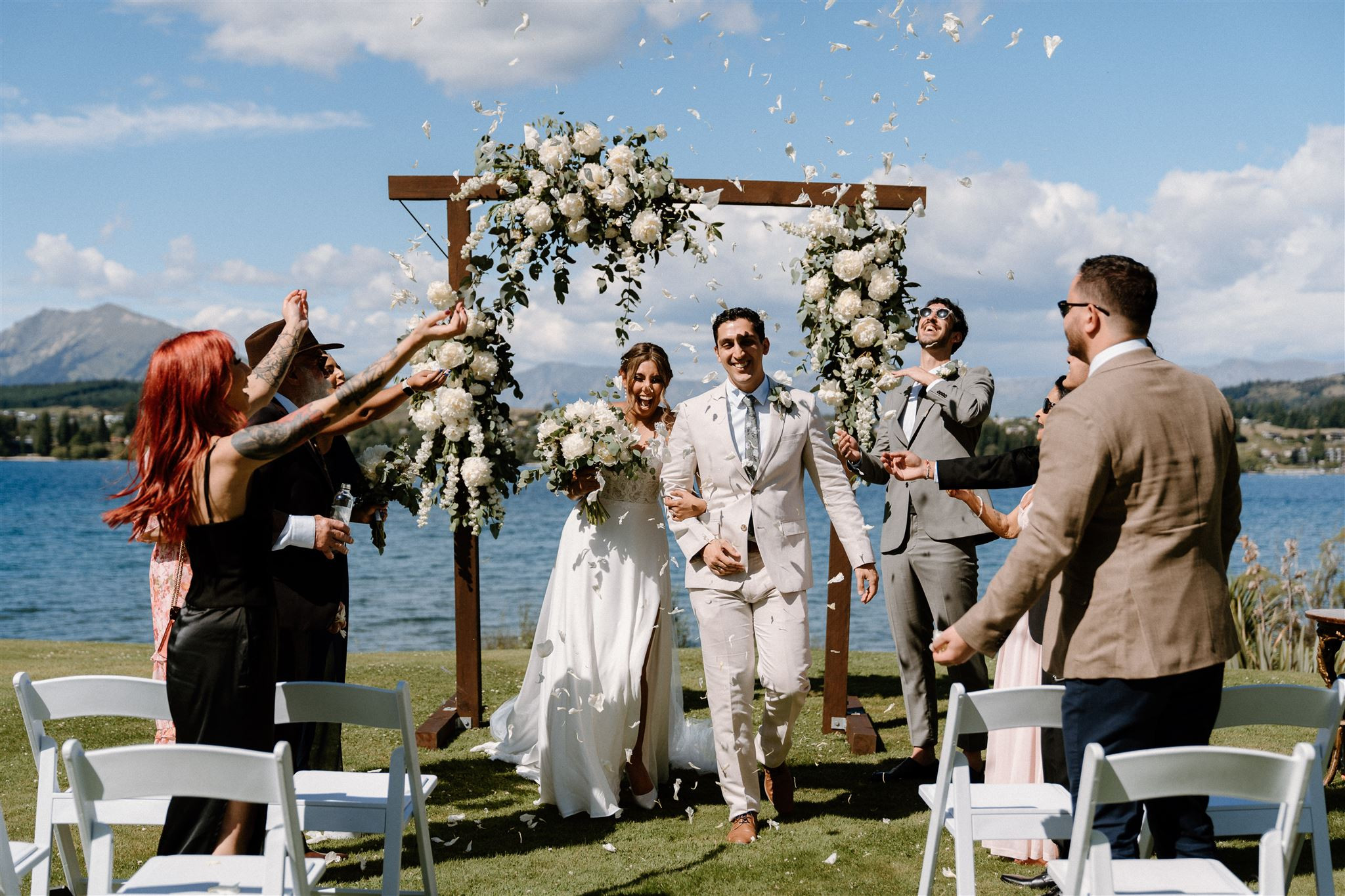 Wedding couple walking down the Aisle with flower petals being thrown above them. 7 guests chaerring them on. Lake Wānaka in the distance