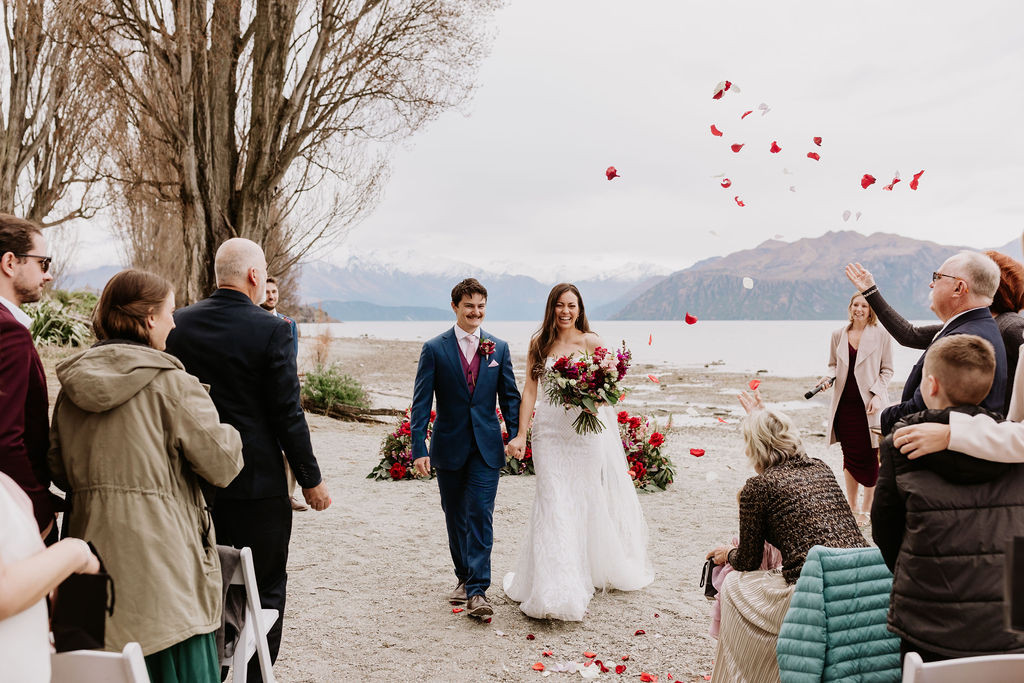 Newly married couple walking down the isle on the Wānaka Lake Front. Flower petals being tossed in the air.  Big Smiles