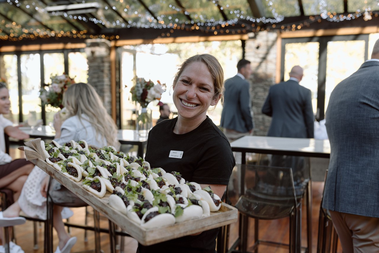 Edgewater Waitress holding a board of Bao Buns, smiling at the camera.  In the background, guests are enjoying the wedding reception